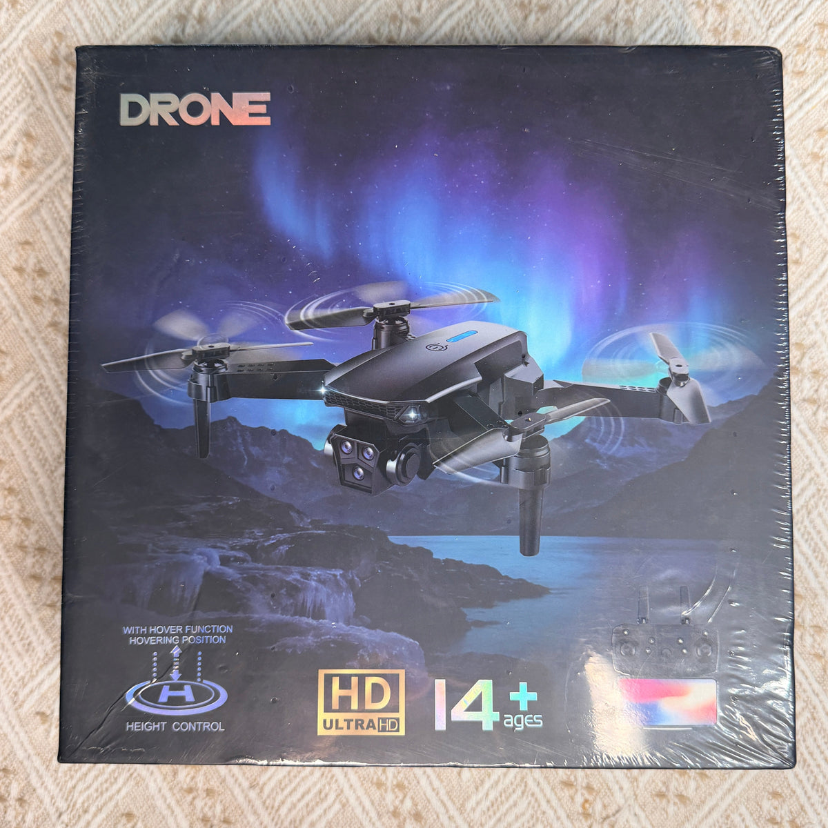 A sealed retail box for a drone, photographed from above on a beige textured fabric surface. The box artwork shows a black quadcopter flying over a rocky landscape with blue aurora lights in the sky.