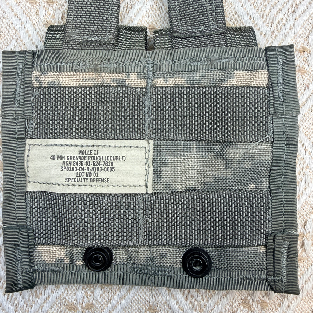 A close-up of a gray MOLLE webbing pouch with a white sewn label reading 'MOLLE IT 40 MM COLLELLA TRUCK (DOUBLE) NSW BARE-50-50C-1005 SPJ030-00-4123-0005 SPECIALTY DEFENSE' and two black snap buttons at the bottom, laid flat on a textured cream fabric background.