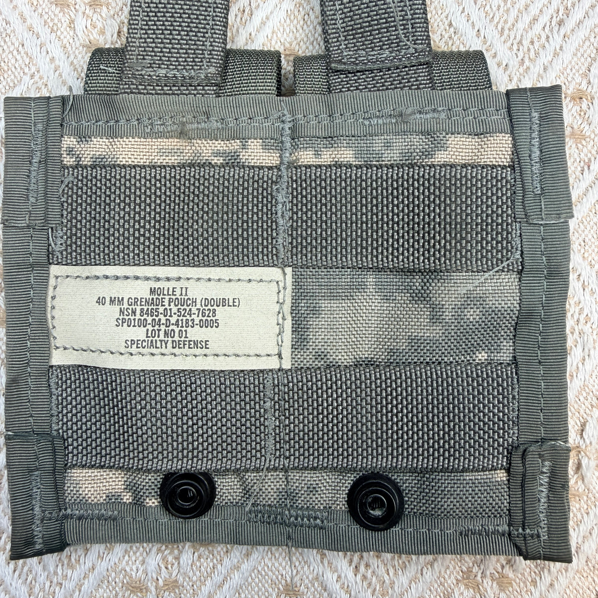 A close-up of a gray MOLLE webbing pouch with a white sewn label reading 'MOLLE IT 40 MM COLLELLA TRUCK (DOUBLE) NSW BARE-50-50C-1005 SPJ030-00-4123-0005 SPECIALTY DEFENSE' and two black snap buttons at the bottom, laid flat on a textured cream fabric background.