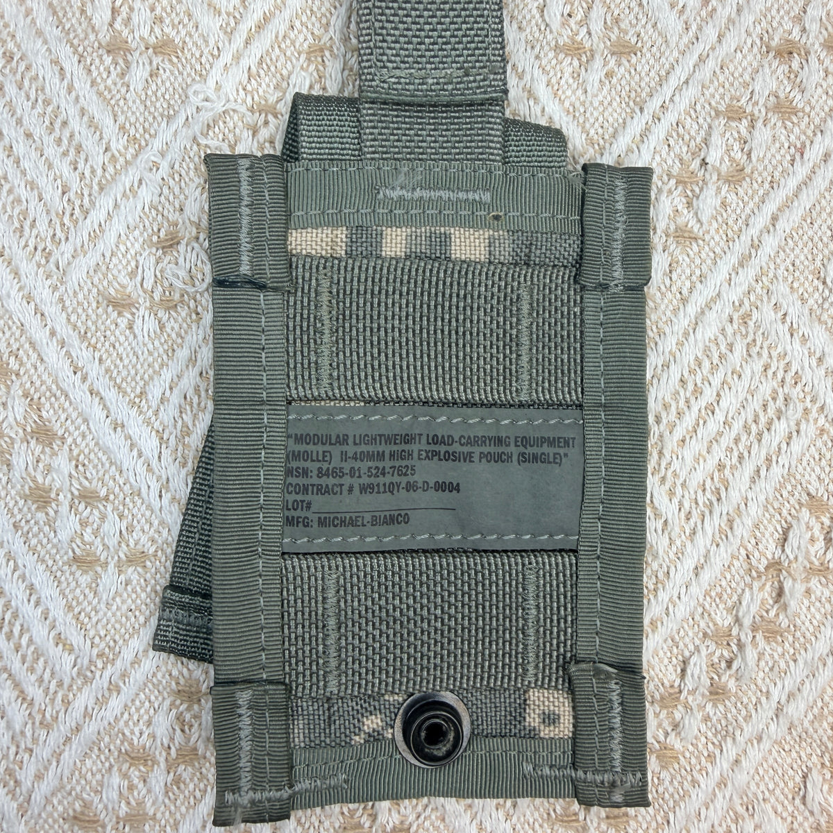 An olive drab modular lightweight load-carrying pouch, model W91207, featuring a black plastic quick-release snap and printed specifications on the front, laid flat on a textured cream fabric background.