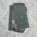 A dark gray tactical utility pouch with a digital camouflage pattern, featuring a snap button closure, laid flat on a textured beige woven fabric background.