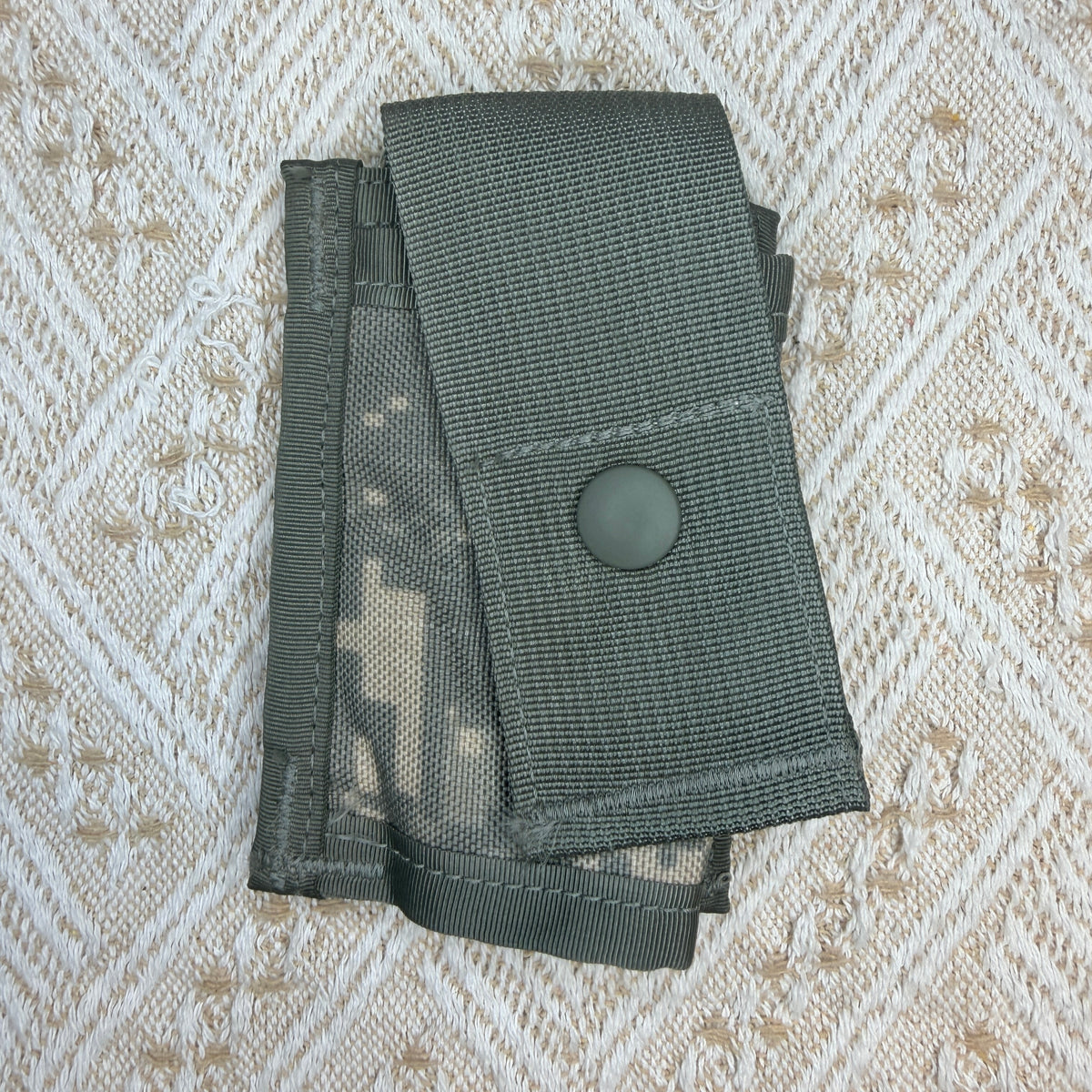 A dark gray tactical utility pouch with a digital camouflage pattern, featuring a snap button closure, laid flat on a textured beige woven fabric background.