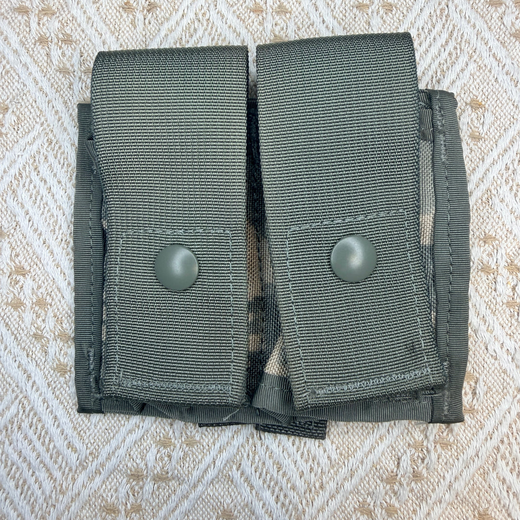 An olive drab green nylon double magazine pouch with two snap-button closures, laid flat on a textured beige and white patterned fabric background.