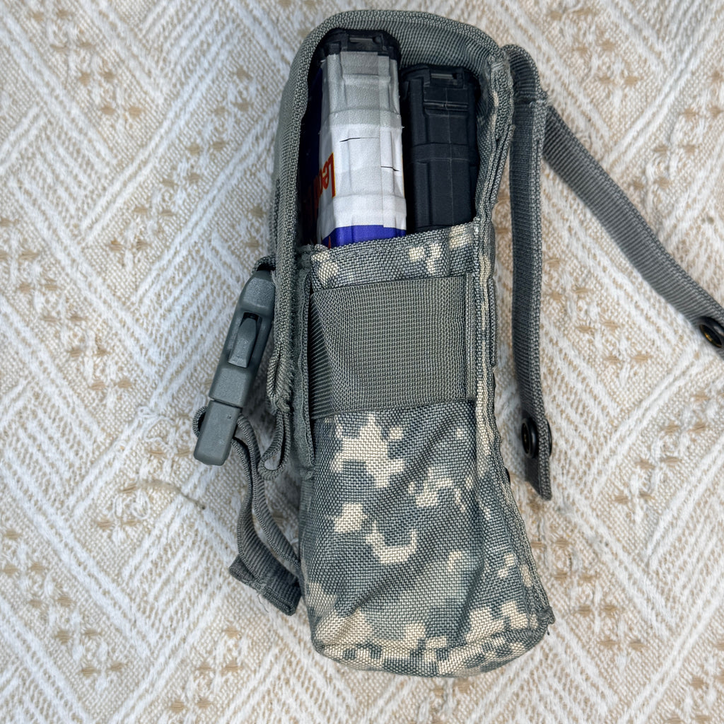 A tactical utility pouch in digital camouflage pattern, shown open with a clear magazine and a black item inside, laid flat on a textured beige and white patterned fabric background.