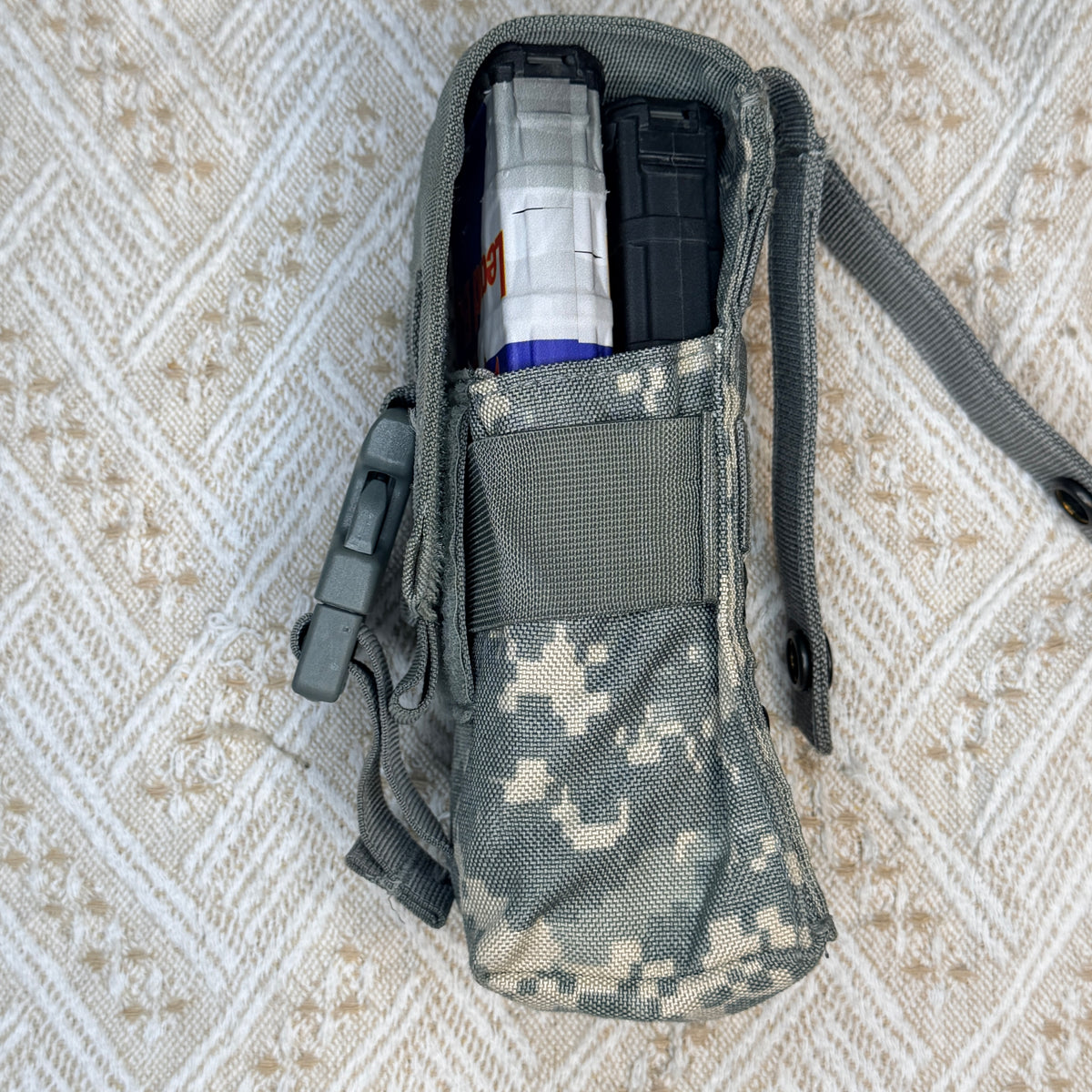 A tactical utility pouch in digital camouflage pattern, shown open with a clear magazine and a black item inside, laid flat on a textured beige and white patterned fabric background.