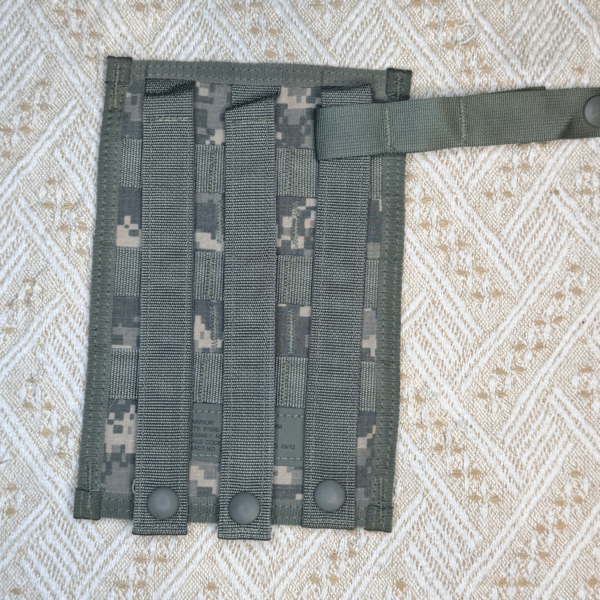 A tactical utility pouch in digital camouflage fabric, featuring four vertical webbing loops with snap buttons at the bottom and a horizontal strap on the right side, laid flat on a textured beige and white woven blanket.