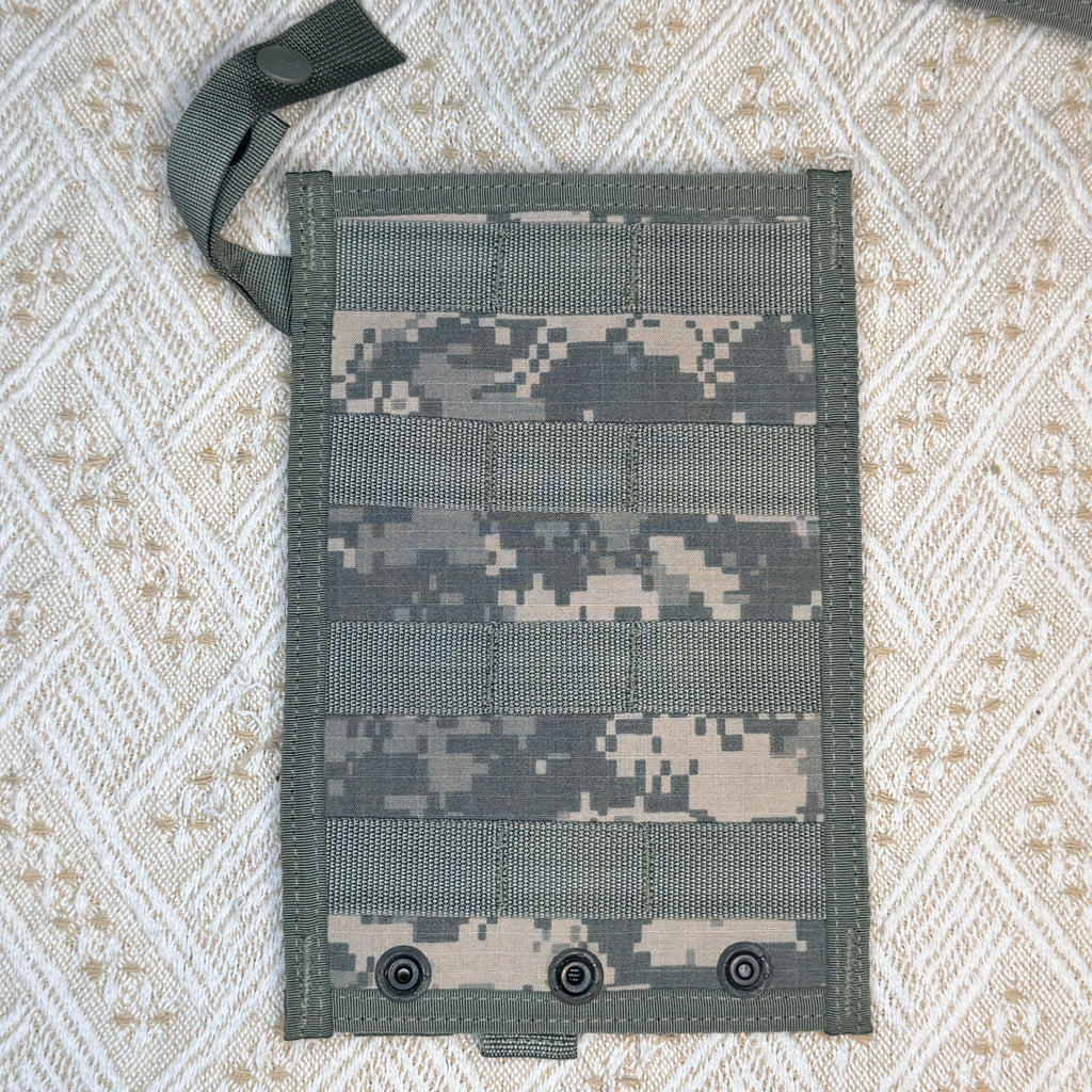 A rectangular tactical webbing pouch with a digital camouflage pattern, featuring three metal snap hooks at the bottom and a matching webbing strap with a snap closure at the top left, laid flat on a textured beige and white woven blanket.