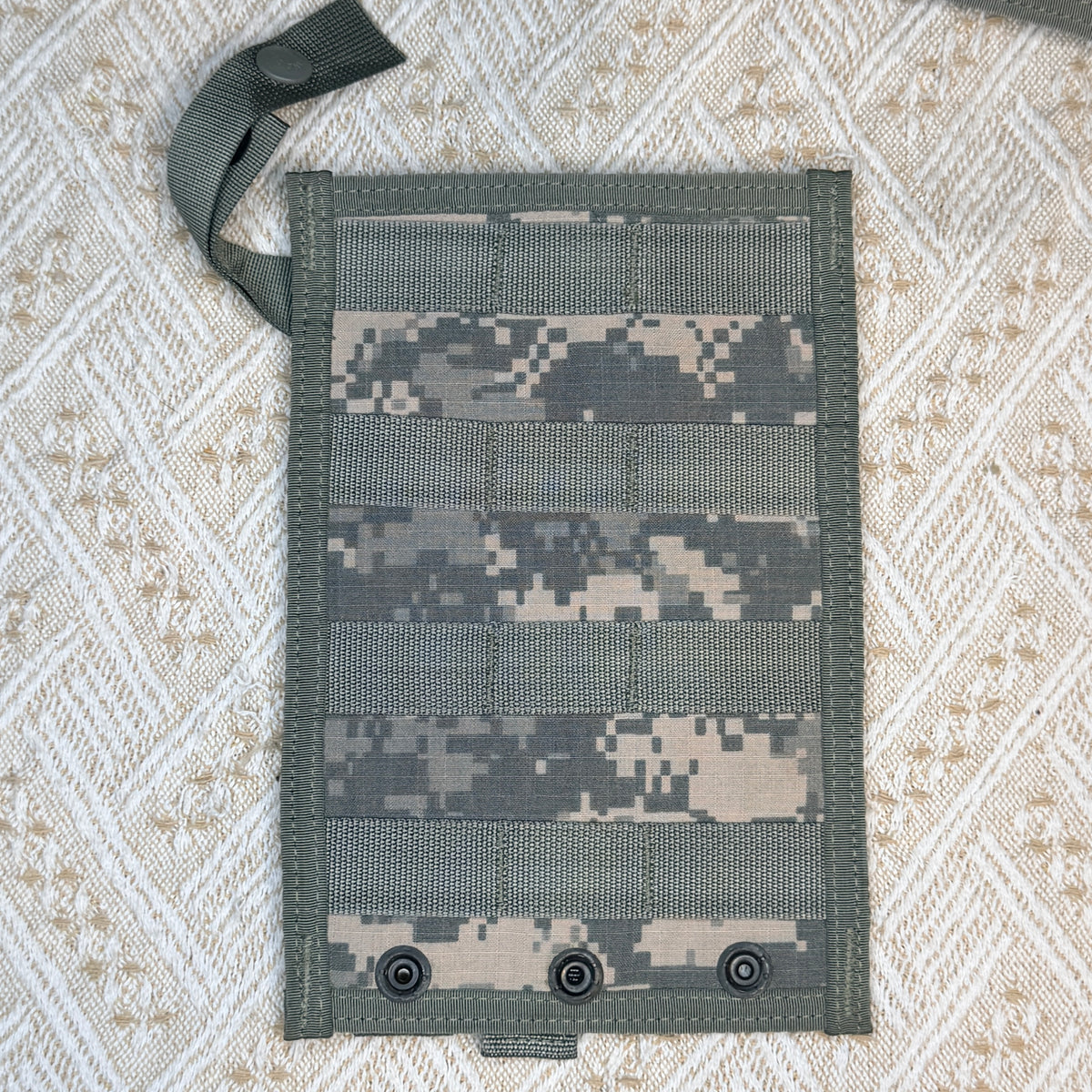 A rectangular tactical webbing pouch with a digital camouflage pattern, featuring three metal snap hooks at the bottom and a matching webbing strap with a snap closure at the top left, laid flat on a textured beige and white woven blanket.