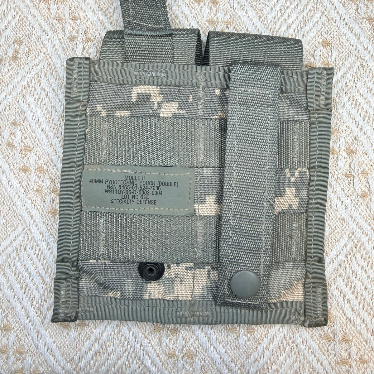 A double 40mm protective pouch in digital camouflage pattern, laid flat on a textured beige and white woven fabric background. The pouch features a hook-and-loop strap closure with a snap button and a sewn-in label.