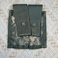 A digital camouflage utility pouch with two front flap pockets secured by dark gray snap buttons, laid flat on a textured beige and white patterned fabric background.