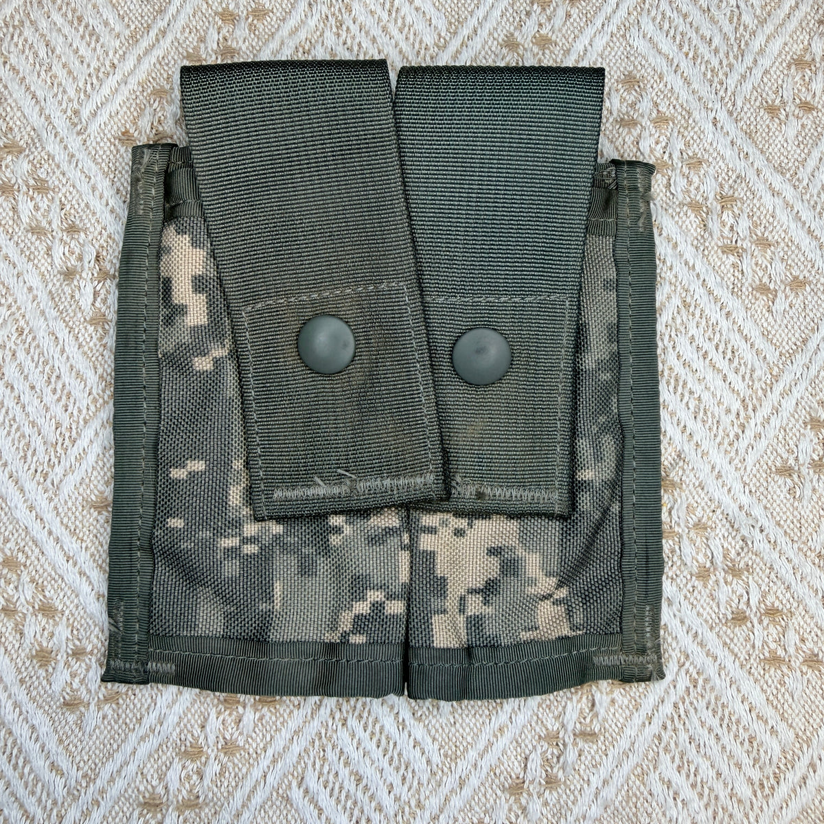 A digital camouflage utility pouch with two front flap pockets secured by dark gray snap buttons, laid flat on a textured beige and white patterned fabric background.