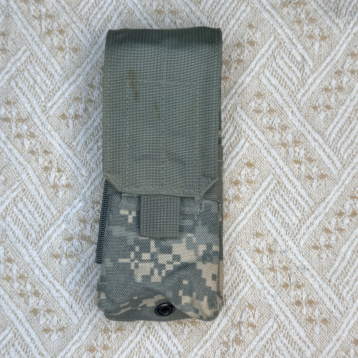 A tactical utility pouch in olive drab and digital camouflage fabric, laid flat on a textured beige and white woven blanket.