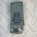 A tactical utility pouch in digital camouflage pattern with a black plastic quick-release buckle, laid flat on a textured beige and white patterned fabric background.
