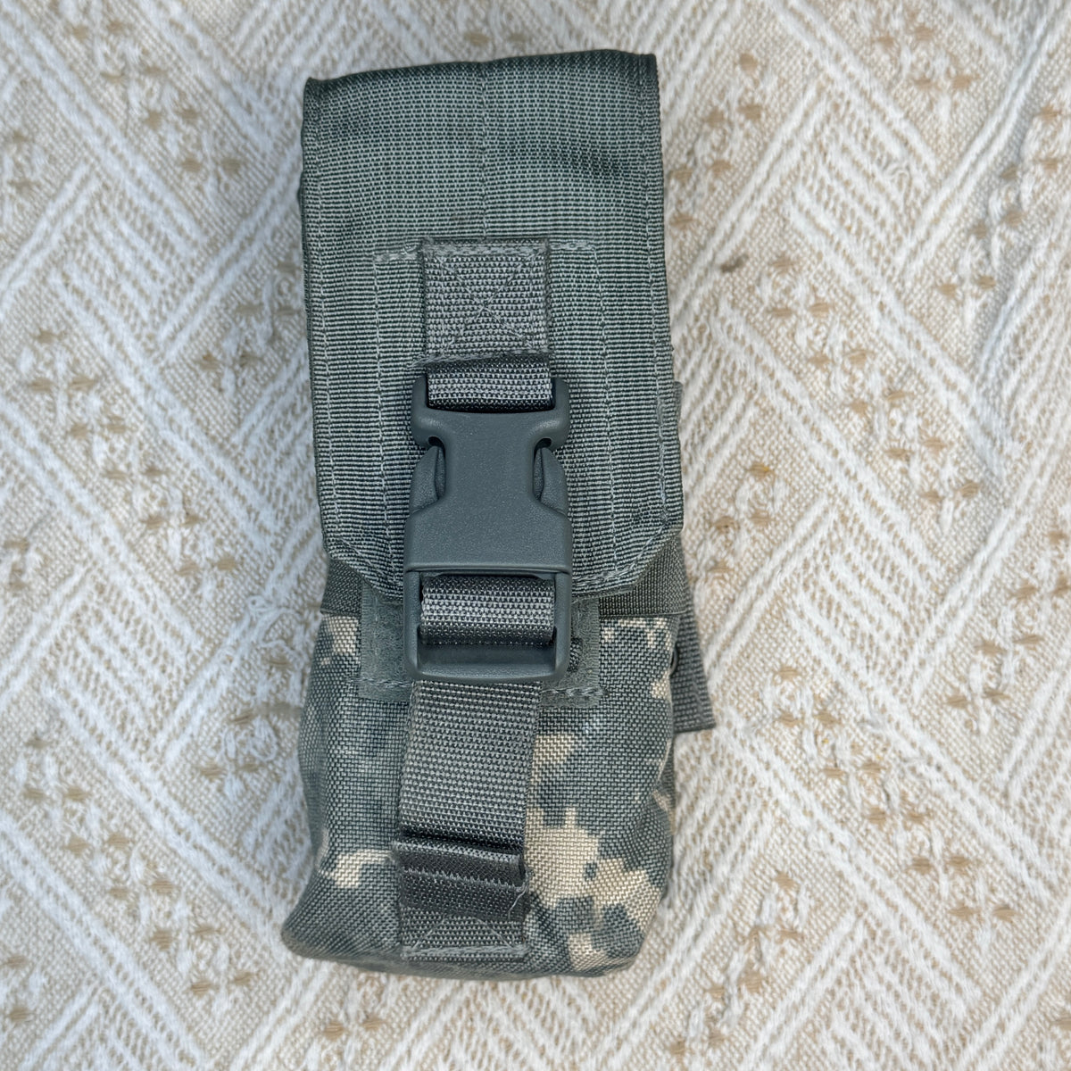 A tactical utility pouch in digital camouflage pattern with a black plastic quick-release buckle, laid flat on a textured beige and white patterned fabric background.
