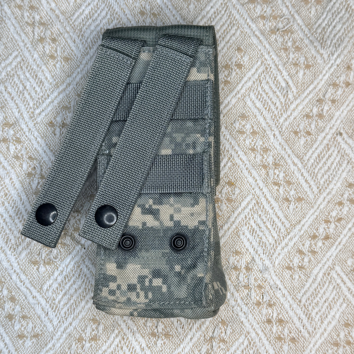 A tactical magazine pouch in digital camouflage pattern, featuring two adjustable webbing straps with metal grommets, laid flat on a textured beige and white woven fabric background.