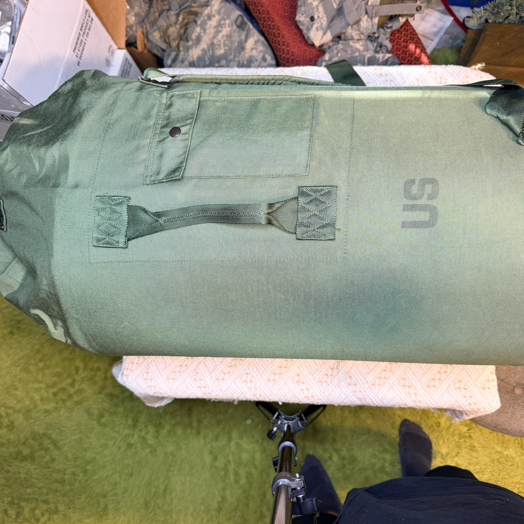 An olive green military-style duffel bag with a top load, a front utility pocket, and a carrying handle.