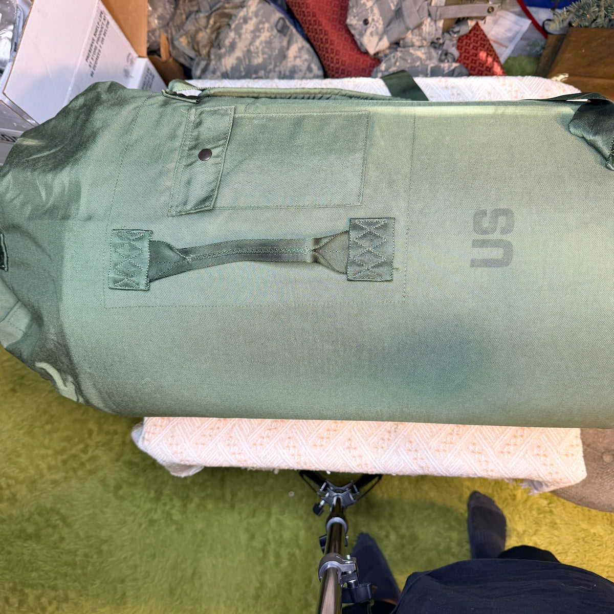 An olive green military-style duffel bag with a top load, a front utility pocket, and a carrying handle.