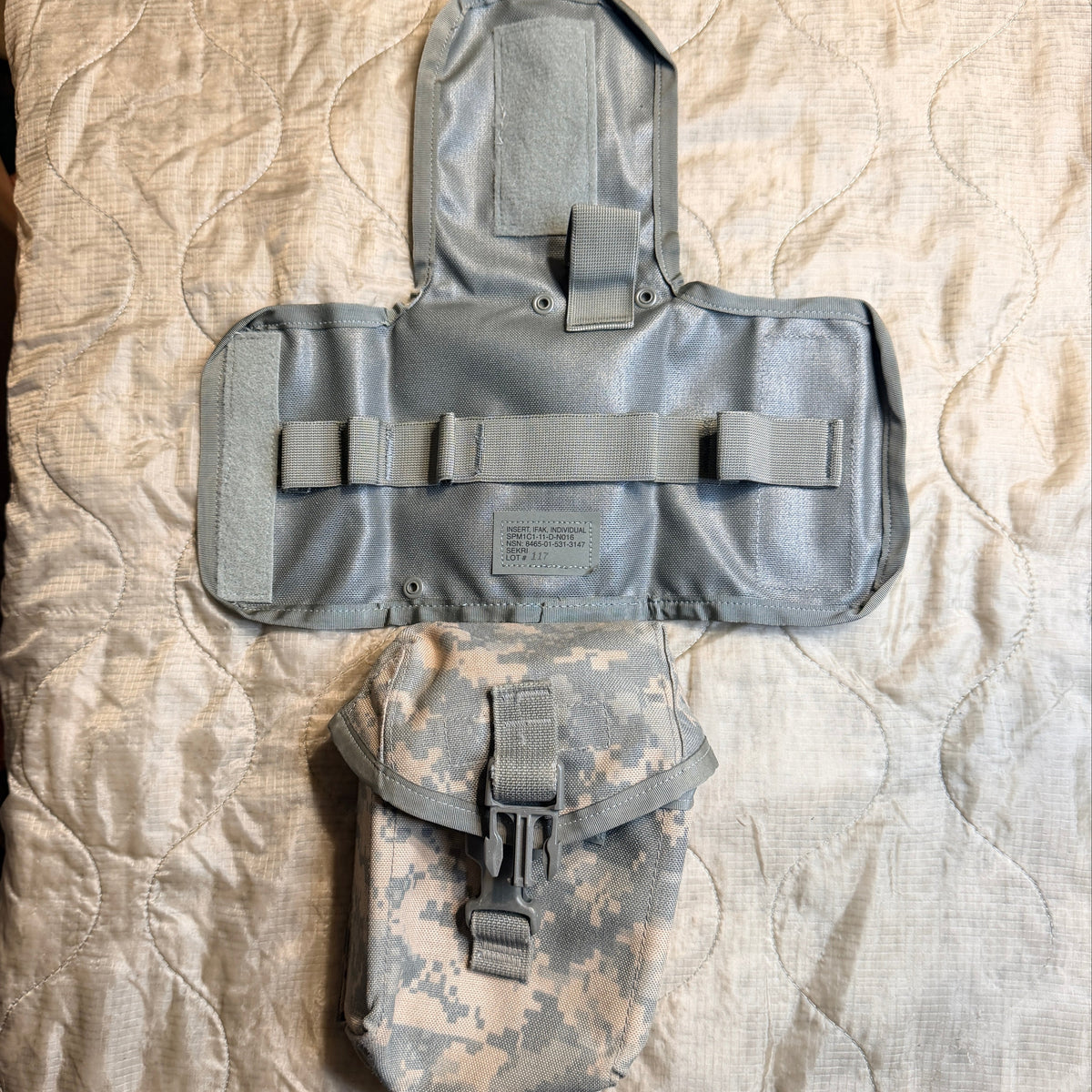 A military utility pouch set laid flat on a quilted bedspread. The set includes a large light gray tactical pouch with a flap and multiple straps, and a smaller digital camouflage pouch below it.