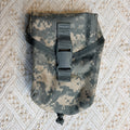 A small tactical pouch in digital camouflage fabric with a black plastic buckle closure, laid flat on a textured beige and white patterned fabric background.