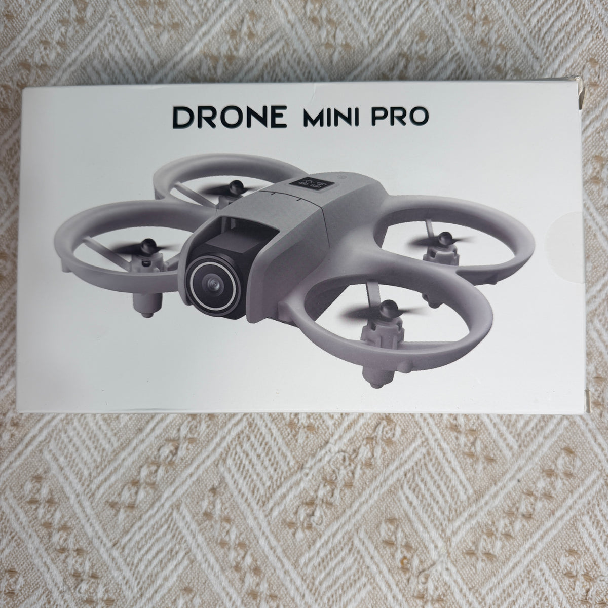 A white product box for a drone mini pro, featuring a printed gray drone illustration, placed on a beige patterned fabric background.