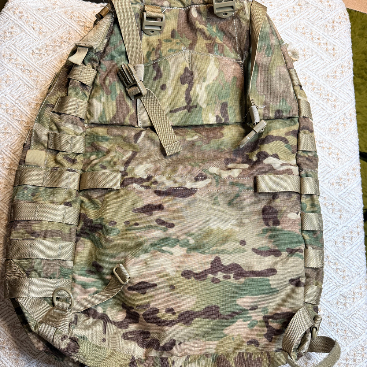 A tactical backpack in a MultiCam camouflage pattern, laid flat on a patterned white fabric surface.
