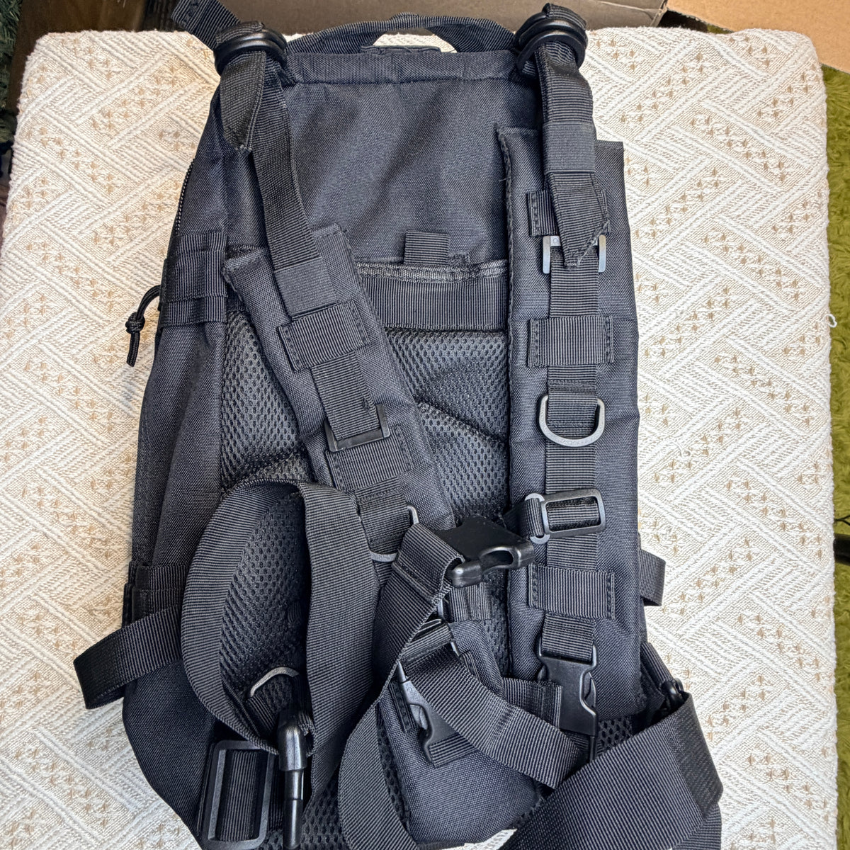 A black tactical backpack shown from the back, featuring multiple adjustable straps, mesh ventilation panels, and plastic buckles, laid flat on a textured beige cushion.