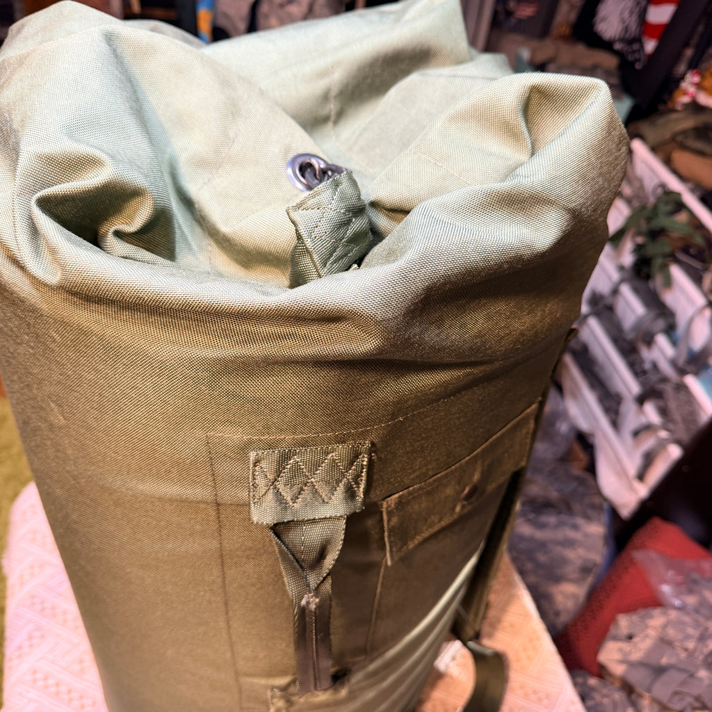 Close-up of a roll-top canvas Duffle Bag in olive green, showing the fabric texture, a metal grommet, and a stitched strap detail.
