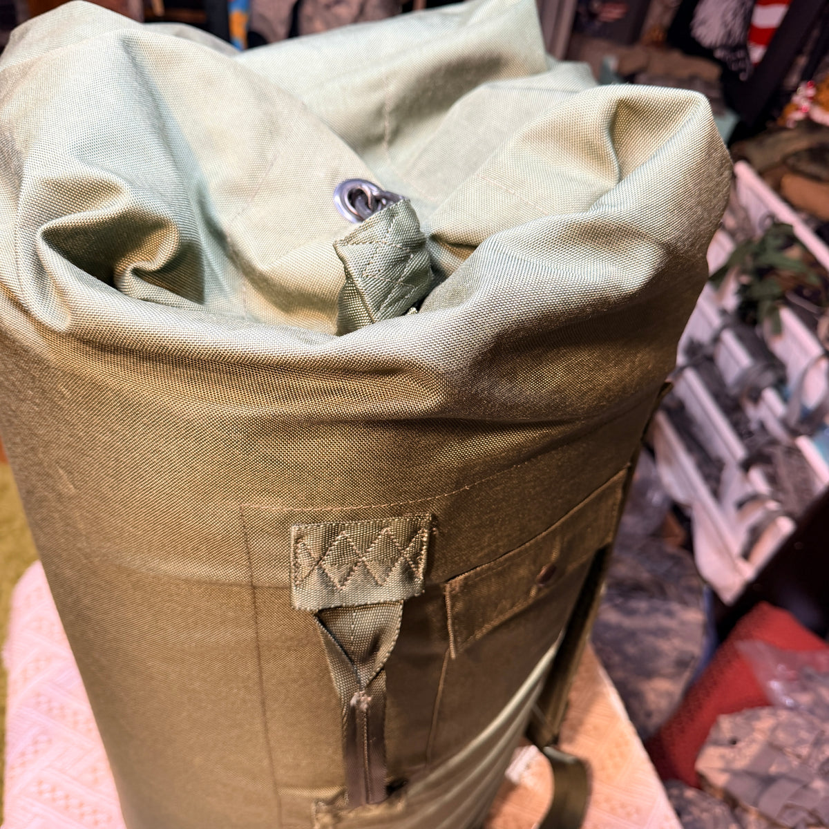 Close-up of a roll-top canvas Duffle Bag in olive green, showing the fabric texture, a metal grommet, and a stitched strap detail.